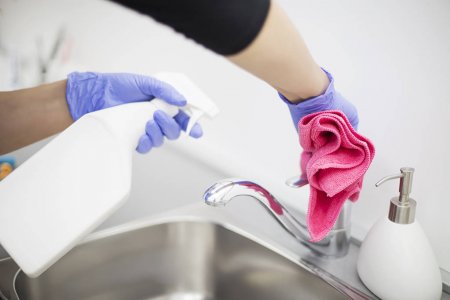 gloved hands with spray bottle and cloth wiping down faucet and sink
